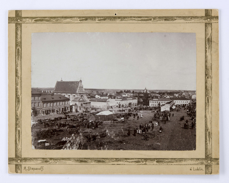 Widok placu targowego przy ulicy Świętoduskiej. Ujęcie od strony odcinka dawnej ulicy Nowej (dzisiejszej Lubartowskiej). Po lewej stronie widoczny kościół karmelitów oraz zabudowa kamienic. Pośrodku plac wypełniony ludźmi i konnymi furmankami. W centralnej części prowizoryczny budynek ze spadzistym jasnym dachem. W głębi po prawej murowany pawilon z kramami. W oddali widoczny kościół i szpital Jana Bożego oraz ówczesny folwark Czechówka.
 
Fotografia sepiowana, naklejona na tekturę, otoczona ozdobną ramką w kolorze złotym w formie przecinających się na rogach listew. W l. d. rogu nazwisko autora fotografii, w p. d. nazwa miasta. Na rewersie odbitki pieczęci tuszowych Muzeum Nałęczowskiego (okrągła, w kolorze fioletowym) oraz Biblioteki Nałęczowskiej (podłużna, w kolorze fioletowym). Przy górnej krawędzi napis informacyjny czarnym tuszem.
