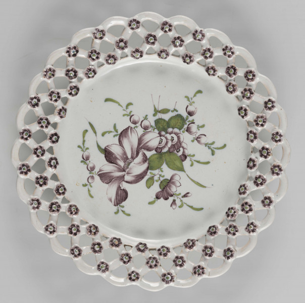 S/CS/1523/ML - Talerz płytki z szerokim ażurowym kołnierzem ze skośnej kratownicy o falistym konturze brzegu. Kratownica zdobiona na złączach kwiatkami. Pokryty polewą w kolorze kremowobiałym; w lustrze duża jasnozielona gałązka z kawiatami w kolorze fioletowej purpury, tego samego koloru kwiatki na kołnierzu.
 