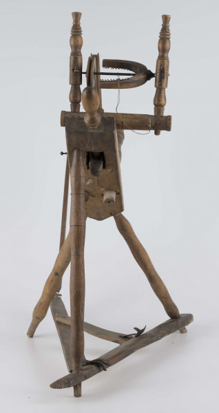 E/16647/ML - Kołowrotek poziomy, drewniany, ozdobnie toczony. Drewno niemalowane. Podstawa to 3 skośnie ustawione nóżki. Dwie z nich osadzone w wąskiej listwie. Górne końce nóżek osadzone w prostokątnej pochyłej grubej desce (11 x 32 x 4 cm). Stanowi ona podstawę do osadzenia koła napędowego i szpuli zwijającej nici. W niej zamocowany uchwyt połączony ze śrubą, która reguluje położenie szpuli. Koło osadzone na metalowej ośce posiada 8 toczonych ozdobnie szprych. Jeden koniec ośki połączony pionową ruchomą listewką z pedałem wprawiającym w ruch kołowrotek. Szpula osadzona również na metalowej ośce.