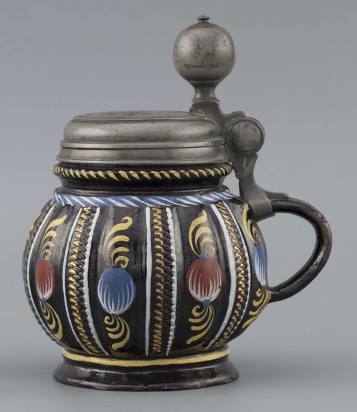 S/CS/595/ML - Kufel  kulisty, o niskiej konicznej stopie i niskiej szyi z pierścieniem, ucho taśmowe w kształcie „C”, pokrywka cynowa. Korpus pionowo kanelowany, podzielony na pasy na przemian gładkie i rytowane w ukośną kratkę. Pokryty szkliwem w kolorze ciemnego kobaltu, zdobiony malowanymi ornamentami podkreślającymi podziały powierzchni, w kolorach: czerwień, żółcień, błękit, biel. Pokrywka płaska, z pierścieniami i uchwytem kulkowym, na zawiasie umocowanym do ucha. Na wierzchu pokrywki grawerunek przedstawiający zawiły ornament – być może monogram spleciony z liter JGL – pod koroną, w uproszczonym wieńcu, z datą 1716.  