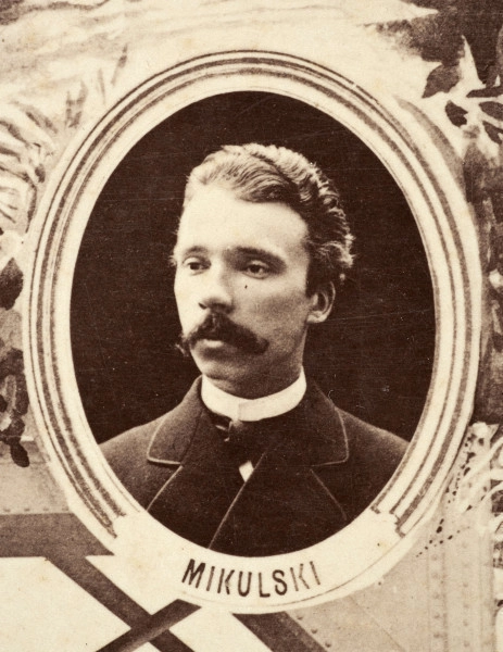 Fotografie w odcieniu sepii umieszczone na tle drzew i roślin, w centralnym miejscu postać kobiety ze sztandarem i aniołkami, wkomponowane widoki budowanej stacji, mostu kolejowego i lokomotywy. Największe zdjęcie w górnej, środkowej części bez podpisu, prawdopodobnie inż. głównej kolei nadwiślańskiej Tadeusz Chrzanowski lub jego następca Walery Titkow. Wśród podobizn pracownicy Biura Głównego Inżyniera: inż. Włodzimierz Stanisławski - pomocnik głównego inżyniera; inż. Feliks Kucharzewski -  pomocnik naczelnika biura; Feliks Kamocki - naczelnik kancelarii; Witold Lanci - naczelnik Wydz. Architektury; dr Wacław Lasocki- służba zdrowia; Władysław Radwan - sprawy zakupu gruntów; inż. Stanisław hr Scipio - projektant mostów kolejowych i jego współpracownicy: inż. Stefan Zieliński, inż. Maurycy Machalski, inż. Feliks Kucharzewski. Fotografia w passe - partout, za szkłem, oprawiona w drewnianą, profilowaną ramę.
