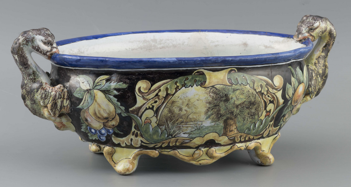 S/CS/90/ML - Żardiniera owalna, na czterech konsolkowych nóżkach połączonych falistymi spływami, wylew z lekko rozchylonym brzegiem, po bokach dwa uchwyty w formie uskrzydlonych smoków, pionowo osadzone. Powierzchni malowana wielobarwnie:  po obu stronach bogate kartusze wolutowo-liściaste z pejzażami  -  jeden z zamkiem obronnym, drugi z wierzbą, po bokach kartuszy festony owoców na ciemno granatowym tle. Wylew obwiedziony błękitnym pasem, wnętrze białobłękitne. 