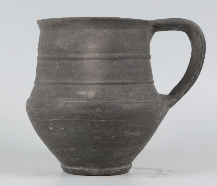 Dno płaskie, wyraźnie wyodrębnione stopką skośnie ściętą ku dołowi; dno wyodrębnione od części przydennej dookolnym wgłębieniem; lekko rozszerzające się ku górze, brzusiec łagodnie zaokraglony, ku górze zwężający się, przechodzący w wysoką, cylindryczną szyjkę; wylew płasko ścięty, rozchylony na zewnątrz; na największej wydętości brzuśca dookolny żłobek; w miejscu przejścia brzuśca w szyję i w połowie szyi dookolny wypukłe listwy; ucho z taśmy środkiem wklęsłej, biegnące od wylewu do załomu brzuśca, szyja zdobiona różnym ornamentem (trzy pasy) przez wygładzenie; powierzchnia gładzona, wyświecona
wymiary dodatkowe: 17,0 cm (obwód); 8,0 cm (śr. dna); 0,3 cm (wys. stopki); 0,5 cm (wys. szyi)