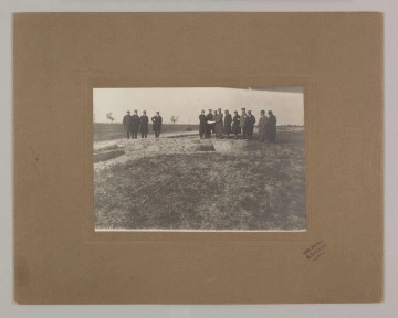 ML/H/F/26/6 - Fotografia przedstawia mężczyzn stojących w dwóch grupach. Po lewej cztery postaci, po prawej dziesięć. Kilku z nich trzyma kartkę papieru. W ziemi widać doły. Fotografia naklejona na brązową tekturę z wytłoczoną ramką. W prawym dolnym rogu pieczęć zakładu fotograficznego S. Zawadzkiego z Lublina.