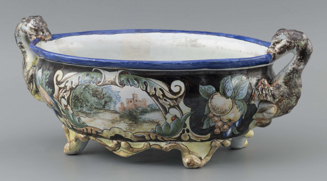 S/CS/90/ML - Żardiniera owalna, na czterech konsolkowych nóżkach połączonych falistymi spływami, wylew z lekko rozchylonym brzegiem, po bokach dwa uchwyty w formie uskrzydlonych smoków, pionowo osadzone. Powierzchni malowana wielobarwnie:  po obu stronach bogate kartusze wolutowo-liściaste z pejzażami  -  jeden z zamkiem obronnym, drugi z wierzbą, po bokach kartuszy festony owoców na ciemno granatowym tle. Wylew obwiedziony błękitnym pasem, wnętrze białobłękitne. 