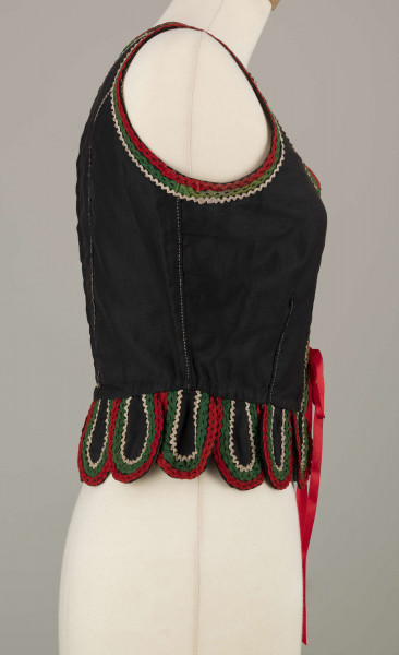 E/16876/ML - Gorset zszyty z 6 kawałków czarnej satyny, podszyty lnianą podszewką. Z przodu rozcięcie usztywnione fiszbinami oraz haftkami do przewlekania wstążeczki. Od pasa dookoła opadają klapki „taszki”. Przód wzdłuż rozcięcie wycięcie wokół szyi, brzegi pach i taszki ozdobione czerwoną, zieloną i białą wełnianą krepinką. Plecy zdobione ornamentem wykonanym ściegiem krzyżykowym. Szyty ręcznie.