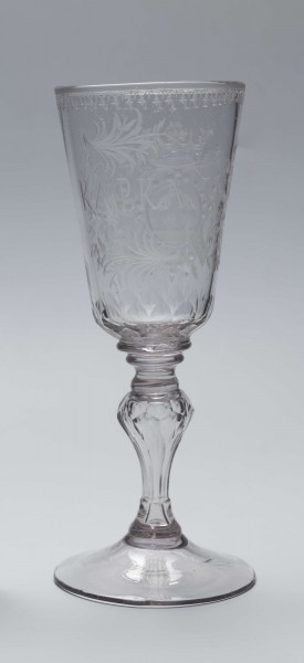 S/CS/244/ML - Stopa okrągła, lekko uniesiona, trzon tralkowy, z pęcherzykiem powietrza, fasetowany, od czaszy oddzielony pierścieniem i dwoma talerzykami, czasza stozkowata, u nasady zaokrąglona, szlifowana w dwa pionowe rzędy owalnych faset. Dekoracja rytowana: na czaszy dwustronna - z jednej strony owalna tarcza z parą gołąbków połączonych dziobkami stojących na serduszkach w rozbudowanym kartuszu z uszakami, z którego wyrastaja gałązki; po drugiej stronie litery 