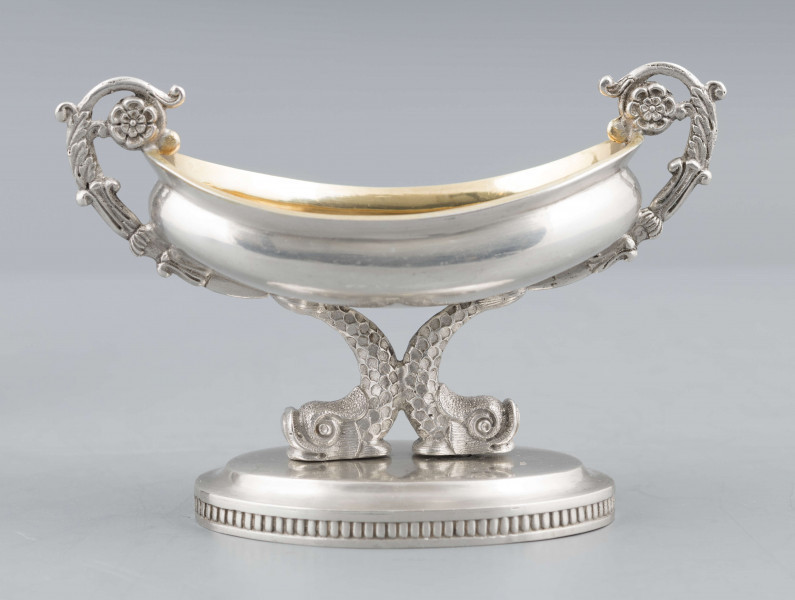 S/Mt/511/ML - Solniczka w formie łódki wspartej na dwóch delfinach, z owalną podstawą, z dwoma uchami z wici roślinnej. Brzeg czarki wywinięty, podstawa lekko podniesiona a jej brzeg zdobiony pionowym karbowaniem. Na spodzie sygnatura:
- imienna 