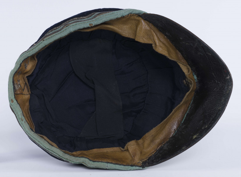 ML/H/2208 - Czapka, czyli tzw. dekiel członka Polskiej Akademickiej Korporacji Astrea działającej na Katolickim Uniwersytecie Lubelskim w latach 1923-1939. Nakrycie głowy w barwach korporanckich, uszyte z nieusztywnianego granatowego sukna, z daszkiem z czarnej skóry, z otokiem w kolorach błękitnym i srebrnym. Na zewnętrznej stronie denka w centralnej części wyszyty fabrycznie srebrną nicią znak korporacji (stylizowany na gwiazdę, w postaci połączonych ze sobą ozdobnych czterech liter „A”) umieszczony wewnątrz haftowanego oktogramu otoczonego kołem z naszytego ozdobnego sznureczka. Po bokach widoczne miejsca zszycia materiału. Wewnątrz podszewka w kolorze granatowym oraz wzmacniający skórzany pasek w kolorze brązowym naokoło.