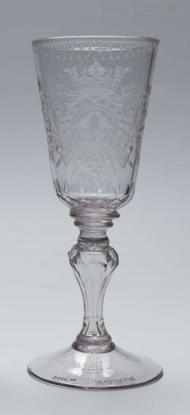 S/CS/244/ML - Stopa okrągła, lekko uniesiona, trzon tralkowy, z pęcherzykiem powietrza, fasetowany, od czaszy oddzielony pierścieniem i dwoma talerzykami, czasza stozkowata, u nasady zaokrąglona, szlifowana w dwa pionowe rzędy owalnych faset. Dekoracja rytowana: na czaszy dwustronna - z jednej strony owalna tarcza z parą gołąbków połączonych dziobkami stojących na serduszkach w rozbudowanym kartuszu z uszakami, z którego wyrastaja gałązki; po drugiej stronie litery 