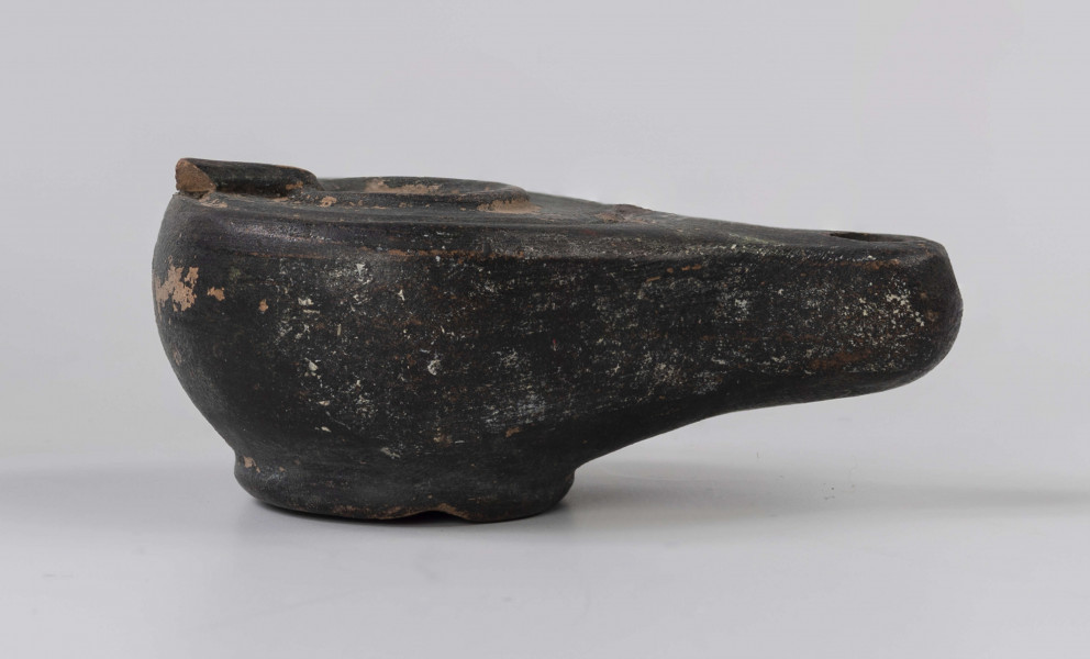 720/A/ML/2 - Lampka oliwna z utłuczonym uszkiem; mały otwór do wlewania oliwy obwiedziony plastycznym wałeczkiem, który jest połączony z uszkiem plastycznym wałeczkiem rozszerzającym się w kierunku uszka; wokół krawędzi dookolny złobek; mały otwór na płomien na wysmukłym, dość mocno wyciągniętym dziubku; dno wyodrębnione, wklęsłe; prawdopodobnie miniatura kultowej, pannońska wzorowana na lampach brązowych