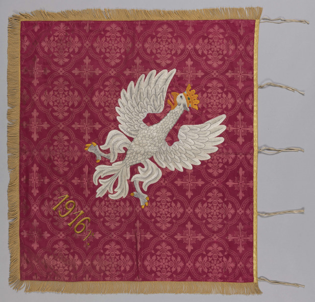 ML/H/200 - Sztandar Przedszkola i Seminarium Ochroniarskiego Marii Papiewskiej. Sztandar w formie kwadratu zszytego z dwóch kawałków ozdobnej tkaniny, obszyty frędzlami. Wzdłuż jednej z krawędzi mocowania w formie troczków do zaczepienia na drzewcu. Na awersie pośrodku tkaniny w kremowym kolorze w kwiatowe motywy herb miasta Lublina (koziołek wsparty o winną latorośl umieszczony na czerwonym polu na tarczy zwieńczonej koroną) oraz haftowany w półkolu czerwoną nicią napis z nazwą posiadacza. Na rewersie pośrodku wzorzystej tkaniny w kolorze czerwonym haftowany biały orzeł w złotej koronie wysadzanej klejnotami z umieszczoną poniżej haftowaną w kolorze złotym datą 1916 r. Metalowa głowica w kształcie orła w koronie umocowanego na kuli. Dodatkowo materiałowy pokrowiec zapinany u góry na napy i haftki, w kolorze zielonkawym, z wyhaftowanym pośrodku napisem w kolorze żółtym.