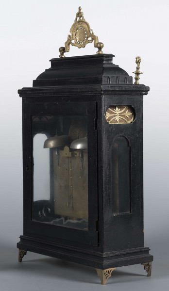 S/Mt/409/ML - Obudowa szafkowa, czworoboczna ze schodkowym zwieńczeniem, pokryta czarną politurą, oszklona z czterech stron, na czterech ażurowych nóżkach z mosiądzu; mosiężny uchwyt, ozdobny, składany a w górnych narożnikach sterczyny w formie płonących wazonów. Mechanizm chodu i bicia o napędzie sprężynowym, z wahadłem i dwoma dzwonkami; bicie godzin i kwadransów, budzik i repetier. Tarcza biało emaliowana z czarnymi cyframi arabskimi godzin i oznaczeniem minut, w niej trzy otwory na osie sprężyn; dwie ozdobne wskazówki. Powyżej tarczy wycięcie z widocznym wahadełkiem kontrolnym i trzy mniejsze emaliowane tarcze: środkowa z sygnaturą: Gottfried / Krohz / Cracovie, boczne z wyłączeniem chodu i bicia.