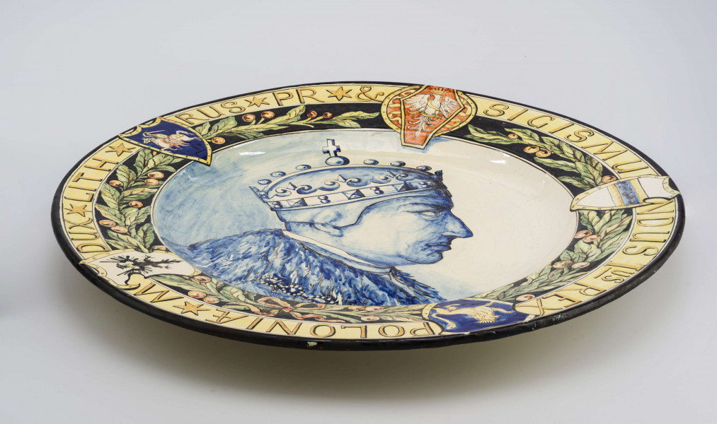 Pólmisek pokrągły, gładki, szkliwiony, dekoracja malowana wielobarwnie. W lustrze malowane błękitem kobaltowym popiersie portretowe Zygmunta I w profilu zwróconym w prawo. Kołnierz podzielony na dwa pasy: od środka zielona girlanda lauru z owocami przewiązana wstążką, na ciemnogranatowym tle; po zewnętrznej  na jasnożółtym tle cieniejsze konturowane litery majuskułowe tworzą napis: SIGISMUN/DUS Ius REX / POLONIAE * M /DUX * LITH */ RUS * PR * &. Pasy dekoracji przedziela pięć herbów regularnie rozmieszczonych: Orzeł, herb Habsburgów, herb Rusi, herb Prus i litewska Pogoń - wszystkie w ozdobnych renesansowych kartuszach. Krawędź obwiedziona ciemnogranatowym konturem.
