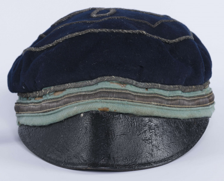 ML/H/2208 - Czapka, czyli tzw. dekiel członka Polskiej Akademickiej Korporacji Astrea działającej na Katolickim Uniwersytecie Lubelskim w latach 1923-1939. Nakrycie głowy w barwach korporanckich, uszyte z nieusztywnianego granatowego sukna, z daszkiem z czarnej skóry, z otokiem w kolorach błękitnym i srebrnym. Na zewnętrznej stronie denka w centralnej części wyszyty fabrycznie srebrną nicią znak korporacji (stylizowany na gwiazdę, w postaci połączonych ze sobą ozdobnych czterech liter „A”) umieszczony wewnątrz haftowanego oktogramu otoczonego kołem z naszytego ozdobnego sznureczka. Po bokach widoczne miejsca zszycia materiału. Wewnątrz podszewka w kolorze granatowym oraz wzmacniający skórzany pasek w kolorze brązowym naokoło.