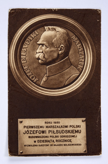 Pocztówka sepiowana - Tablica pamiątkowa ku czci Marszałka Józefa Piłsudskiego wykonana w 10-lecie wojny polsko-bolszewickiej. Ujęcie w pionie.
AWERS: w górnej części karty tondo z wygrawerowanym (?) wizerunkiem Marszałka Józefa Piłsudskiego (ujęcie z profilu w popiersiu), wokół postaci napis; w dole karty przefotografowana pamiątkowa tablica informacyjna (przymocowana w rogach).
REWERS: brązowa linatura z miejscem na korespondencje i znaczek pocztowy (o wartości 15 groszy); przy górnej krawędzi drukiem napis 