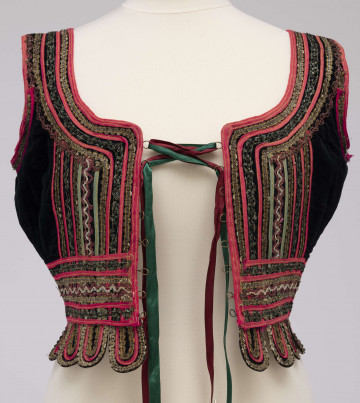 Gorset zszyty z 3 kawałków czarnego aksamitu, podszyty bawełnianą podszewką. Z przodu rozcięcie usztywnione fiszbinami, zapinane na haftki. Od pasa dookoła opadające klapki. Przód, wycięcie wokół szyi, plecy, pas i klapki ozdobione złotym i srebrnym szychem, drobnymi cekinami i wełnianymi tasiemkami w kolorze amarantowym i zielonym. Plecy i boki zdobione żółtą i czerwoną stebnówką. Szyty na maszynie.