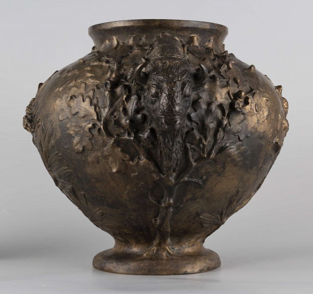 S/Mt/427/ML - Waza w kształcie spłaszczonej kuli, z niską, kolistą stopą i krótką szyją o lekko wywiniętym brzegu. Powierzchnia zdobiona reliefem: po bokach dwa uchwyty w formie głów żubrów, wokół nich gałązki dębu, pośrodku korpusu gałązki sosny z szyszkami wyrastające ze stopy. U góry na brzuścu rytowany napis majuskułą 