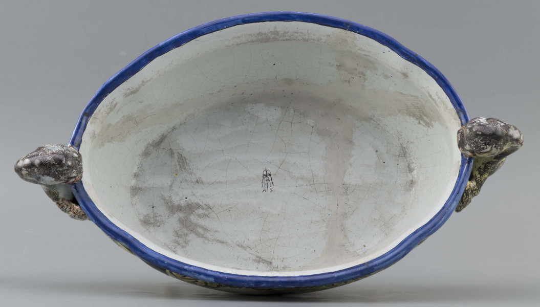 S/CS/90/ML - Żardiniera owalna, na czterech konsolkowych nóżkach połączonych falistymi spływami, wylew z lekko rozchylonym brzegiem, po bokach dwa uchwyty w formie uskrzydlonych smoków, pionowo osadzone. Powierzchni malowana wielobarwnie:  po obu stronach bogate kartusze wolutowo-liściaste z pejzażami  -  jeden z zamkiem obronnym, drugi z wierzbą, po bokach kartuszy festony owoców na ciemno granatowym tle. Wylew obwiedziony błękitnym pasem, wnętrze białobłękitne. 