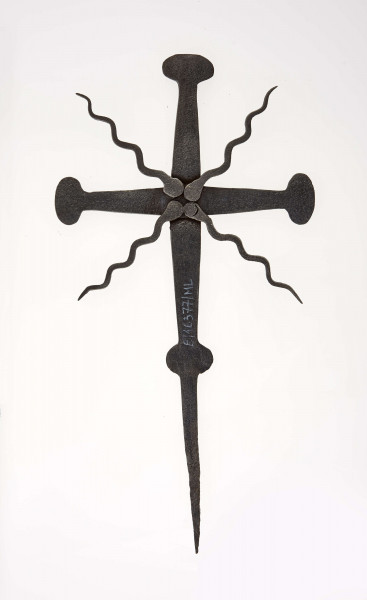 Krzyż żelazny kowalskiej roboty, stanowiący zwieńczenie drewnianego krzyża przydrożnego. Ramiona rozszerzają się ku środkowi, a końce zakończone w kształcie owali. Gr. ramion na końcach 1 cm, przy skrzyżowaniu 2 cm. Między ramionami faliste promienie o dł. 11cm. Szpikulec do wbijania w drewno dł. 13 cm. 