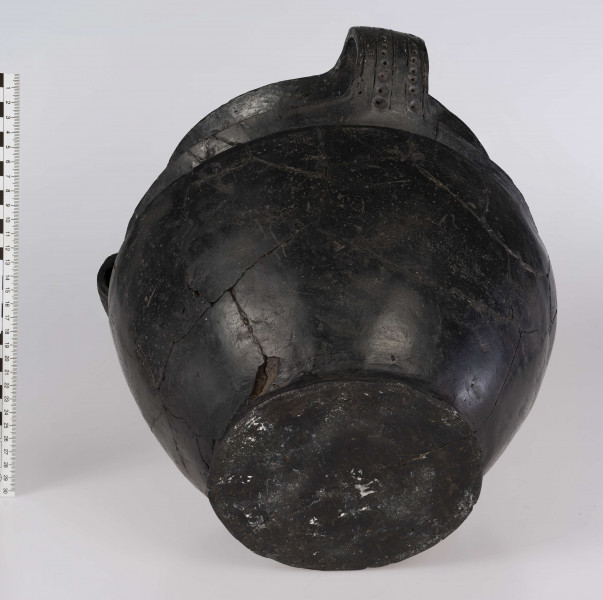 746/A/ML/1 - Waza wielosegmentowa, trzyuszna, wyraźnie profilowana, z odgiętym na zewnątrz prostym brzegiem, ostro zaznaczonym załomem brzuśca i wyodrębionej stopce. Ponad załomem brzuśca znajduje się bogaty, wielowątkowy ornament składający się ze: swastyk tworzonych przez dwie linie ryte, pomiędzy którymi umieszczone są okrąge dołki; z motywu meandra; z kwadratów i prostokątów dzielonych po przekątnej, co tworzy pary przeciwstawnych trójkatów, naprzemianlegle lub promieniście wypełnionych ukośnymi liniami rytymi, niekiedy nakłuciami. Podobnymi ukośnie kreskowanymi trójkątami zdobione są ucha, które posiadają także ornament złożony z pionowych linii rytych, pomiędzy którymi znajdują się różnej wielkości okrągłe dołki wykonane stempelkiem. Barwa czarna.
