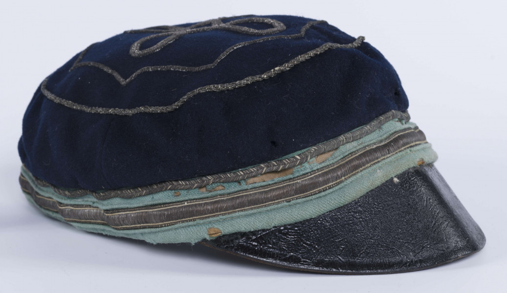 ML/H/2208 - Czapka, czyli tzw. dekiel członka Polskiej Akademickiej Korporacji Astrea działającej na Katolickim Uniwersytecie Lubelskim w latach 1923-1939. Nakrycie głowy w barwach korporanckich, uszyte z nieusztywnianego granatowego sukna, z daszkiem z czarnej skóry, z otokiem w kolorach błękitnym i srebrnym. Na zewnętrznej stronie denka w centralnej części wyszyty fabrycznie srebrną nicią znak korporacji (stylizowany na gwiazdę, w postaci połączonych ze sobą ozdobnych czterech liter „A”) umieszczony wewnątrz haftowanego oktogramu otoczonego kołem z naszytego ozdobnego sznureczka. Po bokach widoczne miejsca zszycia materiału. Wewnątrz podszewka w kolorze granatowym oraz wzmacniający skórzany pasek w kolorze brązowym naokoło.