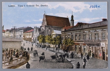 ML/H/3983 - Pocztówka barwna z widokiem na ulicę Świętoduską z sytuowanym przy niej kościołem Karmelitów i targiem. Biegnąca w górę brukowana ulica z szerokim chodnikiem zakręca w lewo. Po prawej stronie chodnik, rząd drzew i jednopiętrowe kamienice. Dalej, na wzniesieniu kościół z renesansowym szczytem. Z lewej fragment zaludnionego targu ogrodzonego drewnianym parkanem oraz ściana murowanych jatek. W głębi słup telegraficzny. W dalekim tle fragment Wieży Wodociągowej na placu Pobernardyńskim (dzisiejszym pl. Wolności). Na ulicy kilka bryczek oraz przechodnie. W l.g. rogu napis informacyjny czarnym nadrukiem. W p.d. rogu data niebieskim długopisem. Na rewersie, pośrodku dolnej krawędzi znak graficzny wydawcy brązowym nadrukiem. Widoczne ślady naklejenia karty na papier. 