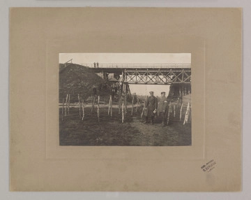 ML/H/F/26/5 ML/H/F/26/5 - Fotografia przedstawia dwóch mężczyzn stojących pośród zasieków nad brzegiem rzeki. Za nimi widoczny most kolejowy i dwie osoby oparte o barierkę mostu. Poniżej kilka osób stoi na skarpie. Fotografia naklejona na szarą tekturę z wytłoczona ramką. W prawym dolnym rogu pieczęć zakładu fotograficznego Stanisława Zawadzkiego z Lublina.
