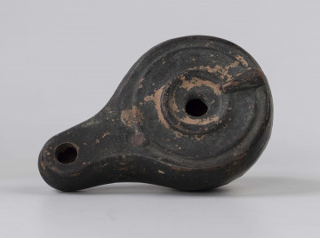 720/A/ML/2 - Lampka oliwna z utłuczonym uszkiem; mały otwór do wlewania oliwy obwiedziony plastycznym wałeczkiem, który jest połączony z uszkiem plastycznym wałeczkiem rozszerzającym się w kierunku uszka; wokół krawędzi dookolny złobek; mały otwór na płomien na wysmukłym, dość mocno wyciągniętym dziubku; dno wyodrębnione, wklęsłe; prawdopodobnie miniatura kultowej, pannońska wzorowana na lampach brązowych