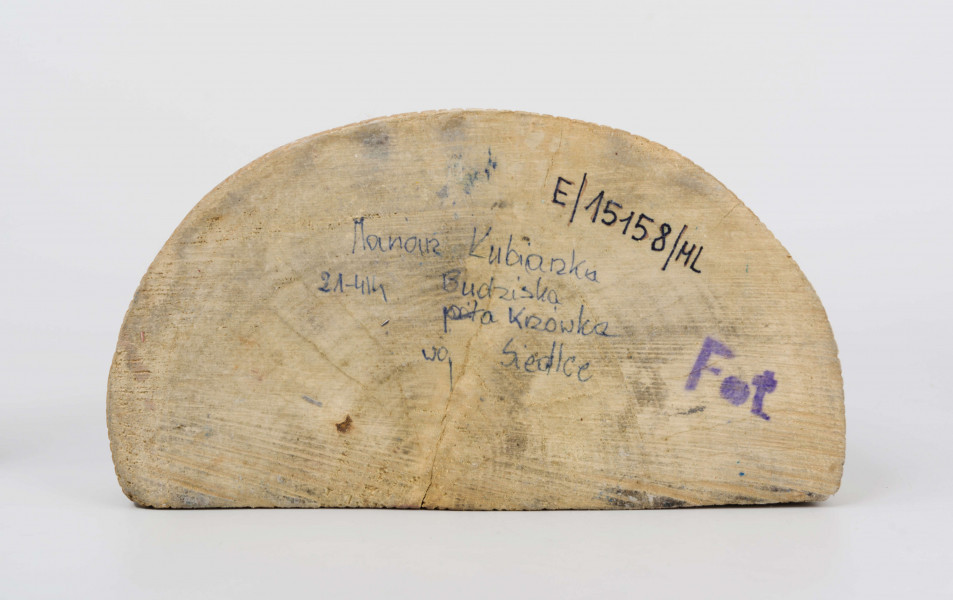 Rzeźba w drewnie lipowym, polichromowana, przedstawiająca postacie matki i dziecka przy kołysce z niemowlęciem. Całość zakomponowana na postumencie wys. 4,4 cm i dł. 22 cm, o kształcie z przodu półokrągłym. Pośrodku usytuowana kołyska na biegunach, wys. 9 cm i dł. 11,2 cm. Kołyska czerwona, ścianki szczytowe ozdobione bukietami składającymi się z trzech wielopłatkowych kwiatów w kolorze żółtym i tulipanowych zielonych liśćmi. W kołysce, w beciku białym, w czerwono-zielone kropki, dziecko, w niebieskiej chustce na głowie. Z lewej strony kołyski postać matki, w długiej jasnoróżowej sukni, w pasie przepasanej fartuchem w żółto-zielono-pomarańczowo-niebieskie poziome pasy. Twarz płaska, nos długi prosty, usta wąskie małe, oczy owalne o podkreślonych białych białkach i czarnych źrenicach, brwi łukowate czarne. Na głowie czerwona chustka zawiązana pod brodą. Spod niej wystają czarne włosy. W rękach trzyma butelkę z mlekiem. Z prawej strony kołyski stoi dziecko w żółtej sukience z niebieskim kołnierzykiem. Twarz dziecka płaska, nos prosty wąski, usta małe, oczy owalne, brwi proste czarne. Włosy czarne, proste sięgające do ramion, zaczesane z grzywką na czole. Ręce wsparte na kołysce. 