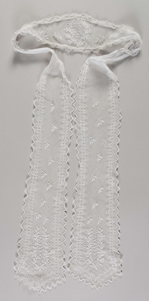 E/16857/ML - Czepek tiulowy biały, składający się z łówki w kształcie diademu oraz 2 rozszerzających się ku końcowi wiązadeł. Na każdym elemencie fabryczne hafty z białej nici. Krawędzie główki obszyte podwójną falbanką z ręcznie wykonanej koronki. Na całej długości głowi hafty z motywem wielopłatkowych kwiatów z łodygami i liśćmi. Pośrodku kwiatostan z pojedynczych pączków otoczony gałązką z liśćmi i pączkami. Krawędzie wiązadeł wycięte w ząbki i wykończone ściegiem obrębiającym. Na powierzchni haft z motywem pojedynczych gałązek z pączkami i liśćmi, w dolnej części – kwiatostan podobny do tego na główce. Wiązadła ozdobione dodatkowo bordiurą  z ząbków.