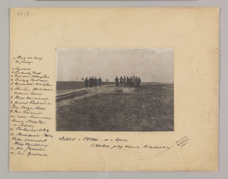 ML/H/F/26/7 - Fotografia przedstawia mężczyzn stojących w dwóch grupach. Po lewej cztery postaci, po prawej dziesięć. Kilku z nich trzyma kartkę papieru. W ziemi widać doły. Fotografia naklejona na kremową tekturę z wytłoczoną ramką. Po lewej stronie odręcznie czarnym atramentem nazwiska postaci z fotografii, pod zdjęciem nazwa miejscowości i data. W prawym dolnym rogu pieczęć zakładu fotograficznego S. Zawadzkiego z Lublina.