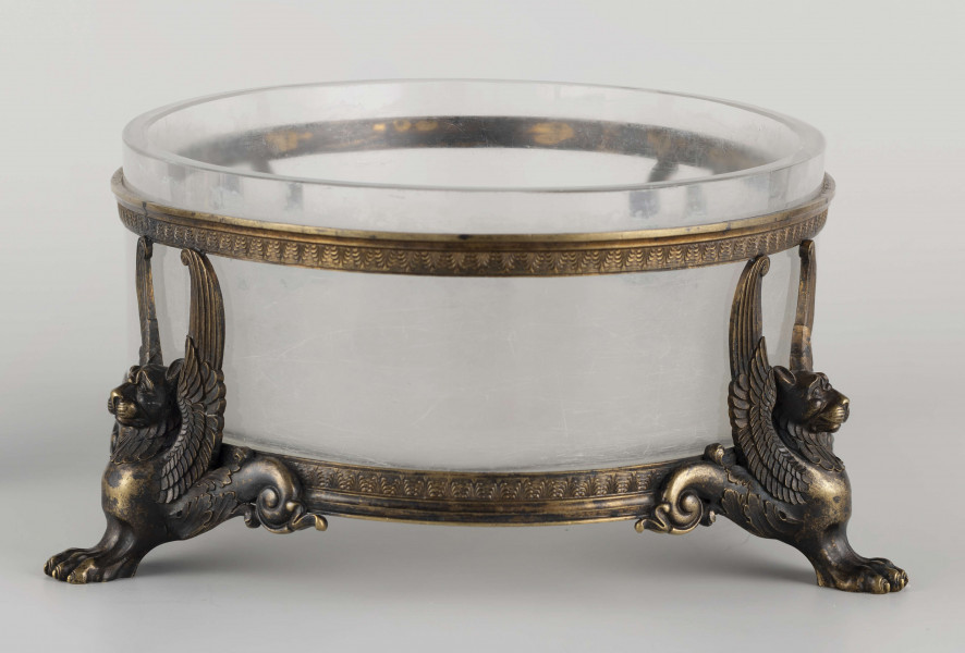 S/CS/1063/ML - Naczynie ze szkła bezbarwnego, grubościennego, owalne, z płaskim dnem, w oprawie metalowej złożonej z dwóch obręczy połączonych plastycznymi figurkami gryfów na nóżkach. Powierzchnia obręczy zdobiona plastycznym fryzem antykizujacych palmet. Oprawa metalowa patynowana. 
 