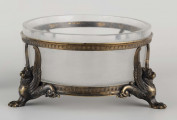 S/CS/1063/ML - Naczynie ze szkła bezbarwnego, grubościennego, owalne, z płaskim dnem, w oprawie metalowej złożonej z dwóch obręczy połączonych plastycznymi figurkami gryfów na nóżkach. Powierzchnia obręczy zdobiona plastycznym fryzem antykizujacych palmet. Oprawa metalowa patynowana. 
 