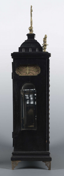 S/Mt/409/ML - Obudowa szafkowa, czworoboczna ze schodkowym zwieńczeniem, pokryta czarną politurą, oszklona z czterech stron, na czterech ażurowych nóżkach z mosiądzu; mosiężny uchwyt, ozdobny, składany a w górnych narożnikach sterczyny w formie płonących wazonów. Mechanizm chodu i bicia o napędzie sprężynowym, z wahadłem i dwoma dzwonkami; bicie godzin i kwadransów, budzik i repetier. Tarcza biało emaliowana z czarnymi cyframi arabskimi godzin i oznaczeniem minut, w niej trzy otwory na osie sprężyn; dwie ozdobne wskazówki. Powyżej tarczy wycięcie z widocznym wahadełkiem kontrolnym i trzy mniejsze emaliowane tarcze: środkowa z sygnaturą: Gottfried / Krohz / Cracovie, boczne z wyłączeniem chodu i bicia.