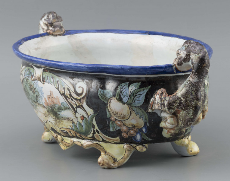 S/CS/90/ML - Żardiniera owalna, na czterech konsolkowych nóżkach połączonych falistymi spływami, wylew z lekko rozchylonym brzegiem, po bokach dwa uchwyty w formie uskrzydlonych smoków, pionowo osadzone. Powierzchni malowana wielobarwnie:  po obu stronach bogate kartusze wolutowo-liściaste z pejzażami  -  jeden z zamkiem obronnym, drugi z wierzbą, po bokach kartuszy festony owoców na ciemno granatowym tle. Wylew obwiedziony błękitnym pasem, wnętrze białobłękitne. 