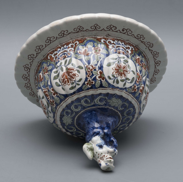 S/CS/546/ML - Wazon owoidalny o niskiej podstawie i niskiej pierścieniowej szyi, powierzchnia drobno kanelowana, pokrywa kopulasta z szerokim kołnierzem, uchwyt w kształcie lwa Fo wspartego prawą łapą na pniu drzewa (?). Dekoracja malowana podszkliwnie w kolorach błękitu, czerwieni, zieleni, żółcieni. W sześciu owalnych, wydłużonych rezerwach na przemian kompozycja kwiatowa z peonią i pejzaż nadrzeczny z architekturą o charakterze orientalnym. U góry - na barkach - sześć owalnych rezerw z kwitnącą gałązką, na dole sześć rezerw trójlistnych z motywem uproszczonej muszli i wolut, na stopie fryz wolutowy, na szyi wąska ukośna kratka. Na pokrywie powtórzona dekoracja z barków, kołnierz biały z fryzem wolutowym analogicznie jak na stopie.
 