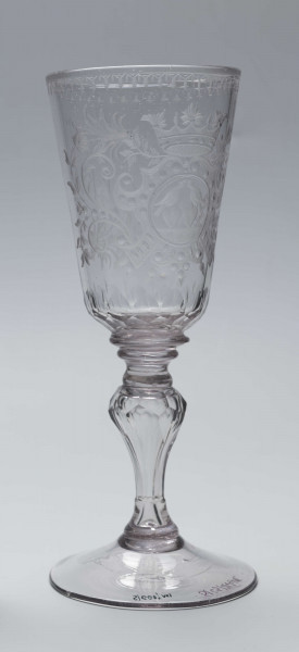 S/CS/244/ML - Stopa okrągła, lekko uniesiona, trzon tralkowy, z pęcherzykiem powietrza, fasetowany, od czaszy oddzielony pierścieniem i dwoma talerzykami, czasza stozkowata, u nasady zaokrąglona, szlifowana w dwa pionowe rzędy owalnych faset. Dekoracja rytowana: na czaszy dwustronna - z jednej strony owalna tarcza z parą gołąbków połączonych dziobkami stojących na serduszkach w rozbudowanym kartuszu z uszakami, z którego wyrastaja gałązki; po drugiej stronie litery 