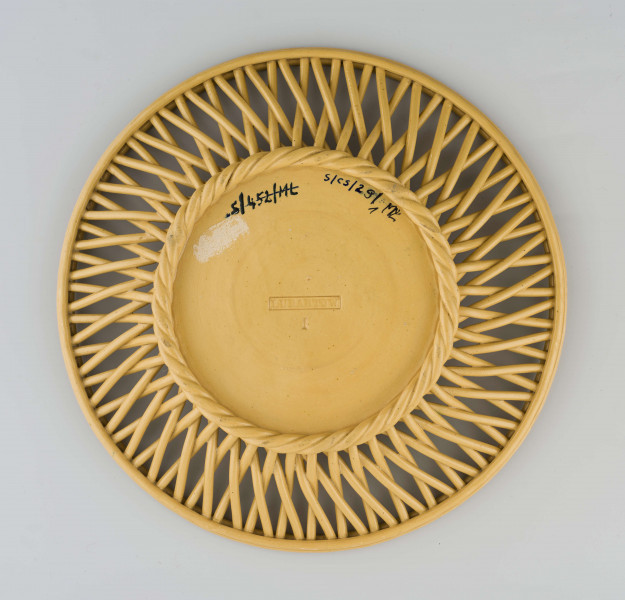  Koszyczek okrągły, płytki; dno płaskie, zdobione plastyczną dekoracją z motywem figuralnym przedstawiającym fauna i amorka pod drzewem; kołnierz ażurowy z okrągłych krzyżujących się wałków połączonych profilowaną obręczą tworzącą krawędź. Całość pokryta połyskliwą glazurą w kolorze ugru.
 