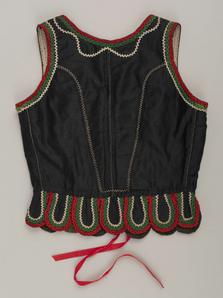 E/16876/ML - Gorset zszyty z 6 kawałków czarnej satyny, podszyty lnianą podszewką. Z przodu rozcięcie usztywnione fiszbinami oraz haftkami do przewlekania wstążeczki. Od pasa dookoła opadają klapki „taszki”. Przód wzdłuż rozcięcie wycięcie wokół szyi, brzegi pach i taszki ozdobione czerwoną, zieloną i białą wełnianą krepinką. Plecy zdobione ornamentem wykonanym ściegiem krzyżykowym. Szyty ręcznie.