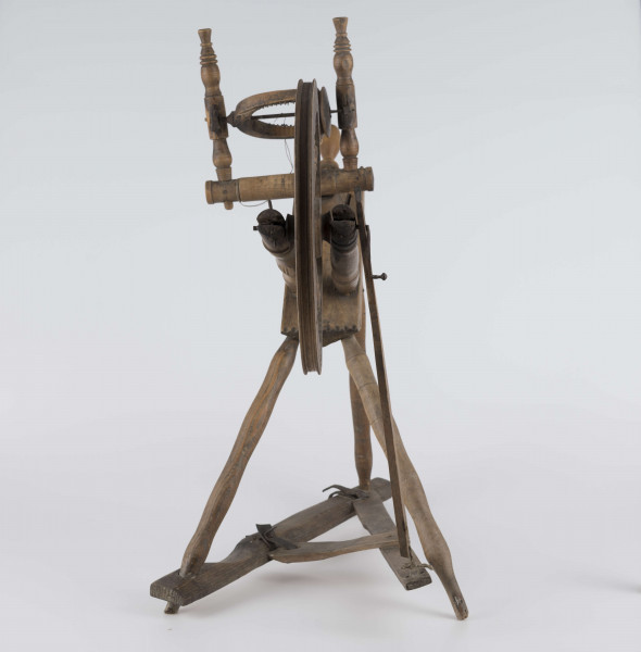 E/16647/ML - Kołowrotek poziomy, drewniany, ozdobnie toczony. Drewno niemalowane. Podstawa to 3 skośnie ustawione nóżki. Dwie z nich osadzone w wąskiej listwie. Górne końce nóżek osadzone w prostokątnej pochyłej grubej desce (11 x 32 x 4 cm). Stanowi ona podstawę do osadzenia koła napędowego i szpuli zwijającej nici. W niej zamocowany uchwyt połączony ze śrubą, która reguluje położenie szpuli. Koło osadzone na metalowej ośce posiada 8 toczonych ozdobnie szprych. Jeden koniec ośki połączony pionową ruchomą listewką z pedałem wprawiającym w ruch kołowrotek. Szpula osadzona również na metalowej ośce.