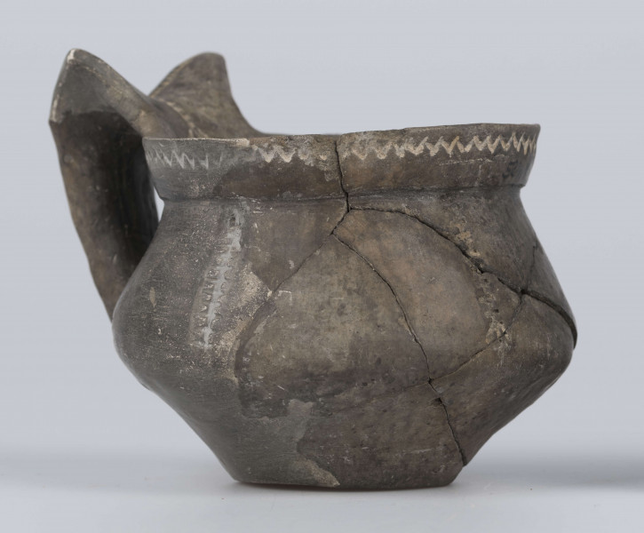 524/A/ML/2 - Kubek gliniany o niskiej szyjce, lejkowato rozchylonej o ukośnie ściętej do wewnątrz krawędzi, oddzielona ostrym załomem od dwustozkowatego brzusca, którego maksymalna wydętość przypada nieco poniżej połowy wysokości naczynia, dno silnie wyodrębnione, wklęsłe. Górny przyczep ucha ansa lunata przylega do krawędzi wylewu, dolny siega największej wydętości brzuśca. Ornament odciśniety prostokątnym stempelkiem, pod wylewem pozioma linia zygzakowata, na brzuścu 6 pionowych pasm ornamentu drabinkowego zakończonych 3 ukośnymi odciskami stempelka (sięgających od załomu szyjka/brzusiec po maksymalną wydętość brzuśca). Takie pasmo także na osi symetrii ucha, pozioma linia drabinkowa łączy skrzydełka ucha anasa lunata. Ornament inkrustowany białą masą. Wymiary: wysokość 7,4 cm; wysokość z uchem ok. 9,1 cm; średnica wylewu 8 cm; szyjka 7,1 cm; brzusiec 9,5 cm; dno 4,4 cm. Surowiec - glina z drobnym piaskiem. Barwa powierzchni zewnętrznej - szarobrunatna, powierzchni wewnętrznej brunatna. Przełom szary. Sposób wykonania - lepienie, powierzchnia gładzona do wybłyszczenia, zdobiona, dobry wypał. zabiegi konserwatorskie - oczyszczanie, sklejanie, wygipsowanie, ucho częściowo zrekonstruowane. Stan zachowania - średni.