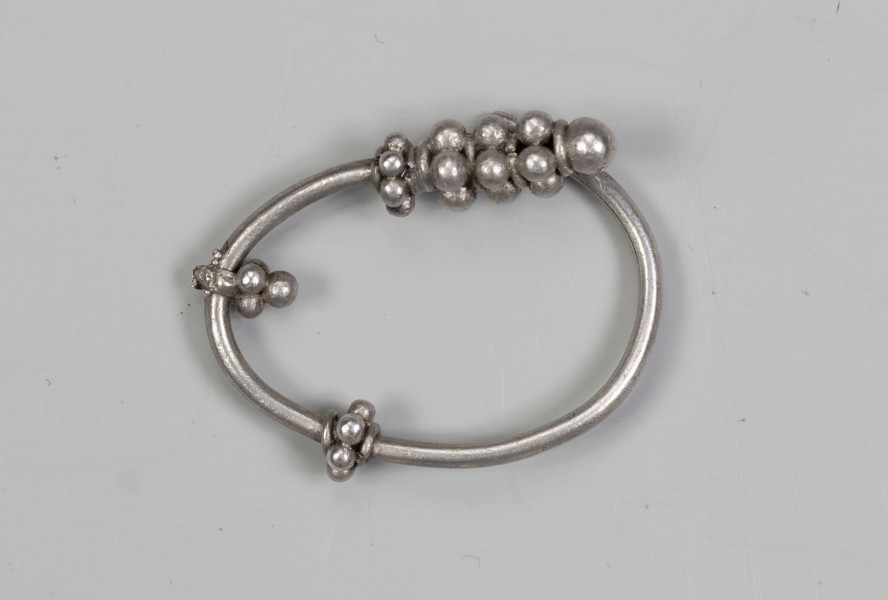 169/A/ML/1 - Zausznica srebrna składa się z kabłąka, na którym zawieszono wisiorek o winogronowatym kształcie. Ozdoby tego typu charakteryzują się kabłąkiem o jednym końcu ucięty prosto, drugim zaś zwiniętym w oczko. W połowie wysokości kabłąka znajdują się wieńce wykonane z granulek ograniczonych pierścieniami z drutu. W dolnej części kabłąka występuje wisiorek uformowany z kilku wieńców granulek zwieńczony granulką osadzoną na pierścieniu z drutu. 