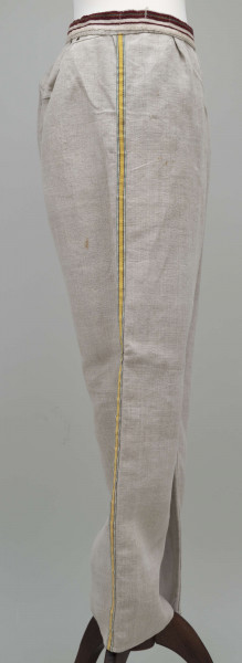 E/12943/ML - Spodnie z lnianego materiału tkanego splotem prostym. Szyte ręcznie i na maszynie. Nogawki szerokie ujęte w pasek szer. 3 cm, wzmocniony i ozdobiony paskiem płótna z ornamentem tkackim „pereborami”. Poniżej paska wszyte 2 trójkątne kliny oraz podobny w rozporku, zapinanym na 2 guziki. Na szwach bocznych nogawek naszyte kolorowe, fabryczne tasiemki, szer. 1,5 cm – żółta i niebieska.