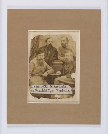 ML/H/565/2 ML/H/565/2 - Powstańcy styczniowi zesłani na Syberię: Julian Kędrzycki z Podola, Mieczysław Bardecki z Żytomierskiego, hr Zygmunt Krasicki z Podola, Z. Rudnicki z Wołynia. Fotografia grupowa czterech mężczyzn w dojrzałym wieku. Dwie osoby stoją z rękoma wspartymi o drewniany cokół. Dwie siedzą z dłońmi opartymi o blat stolika. Postać siedząca po lewej w kożuchu z wykładanym futrzanym kołnierzem, czapką z okrągłym denkiem bez daszka, lewą dłonią wspartą o stół, z długą brodą i twarzą zwróconą w kierunku obiektywu. Postać stojąca po lewej w okularach, półdługich włosach zaczesanych na bok, z brodą przystrzyżoną w szpic, w ciemnych kamizelce i marynarce z wykładanymi kołnierzami. Postać siedząca po prawej z prawą dłonią wspartą o stół, lewą opartą o nogę założoną na kolano, z włosami zaczesanymi na bok i gęstą brodą, ubrana w kamizelkę i marynarkę z wykładanymi kołnierzami. Postać stojąca po prawej w jasnej marynarce ze stójką pod szyją, zarostem na policzkach i brodzie, włosami zaczesanymi do tyłu, twarz lekko bokiem. U dołu zdjęcia na wydłużonym białym polu nazwiska sfotografowanych. Zdjęcie z ubytkami na awersie, podklejone na tekturę.