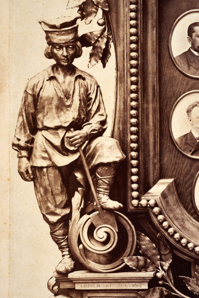 Pośrodku prostokątnego pola o zaokrąglonych narożnikach większa fotografia przyozdobiona wstęgą i gałązkami wawrzynu. Dookoła szesnaście symetrycznie rozmieszczonych mniejszych fotografii. W prawym górnym rogu dr Wacław Lasocki, naczelny lekarz kolei nadwiślańskiej w latach 1875 - 78. W polu ramy w górnej części medalion z płaskorzeźbionym popiersiem z profilu. Przedstawia Leopolda Kronenberga - ojca. Po bokach dwia półleżące aniołki trzymają wieniec laurowy. Po lewej stronie ramy postać robotnika Poleszuka, po prawej pracownika warsztatów kolejowych. W dolnej części front lokomotywy i złoty żeton uprawniający do bezpłatnej jazdy pociągiem I klasy. Fotografia naklejona na tekturę, w dolnej części u dołu sucha pieczęć zakładu fotograficznego.