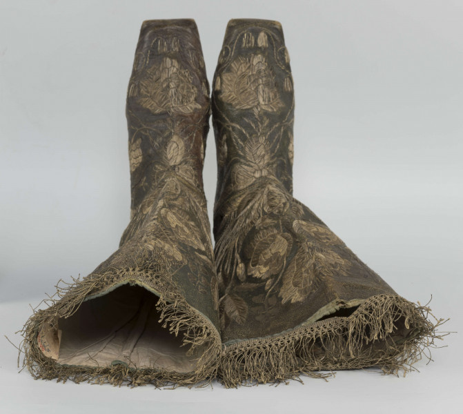 S/T/42/ML - Para butów z wysokimi cholewkami. Cholewka z miękkiej,zielonej skóry,  z dekoracyjnym haftem złotą nicią. Haft z dużym motywem kwiatowym: piwonie, kampanule i drobne kwiatki niezapominajki, tulipany(?), wydłużone liście i wici roślinne, wygięte łukowato.  Wewnatrz buty mają wykończenie białym, cienkim płótnem. Brzegi cholewki obszyte złota koronką z frędzlą. Podeszwy cieńkie z jednego kawałka skóry, bez obcasów, płaskie, z przodu prosto ścięte. Obydwie, skórzane podeszwy mają taki sam wykrój, bez rozróżnienia na lewą i prawą.