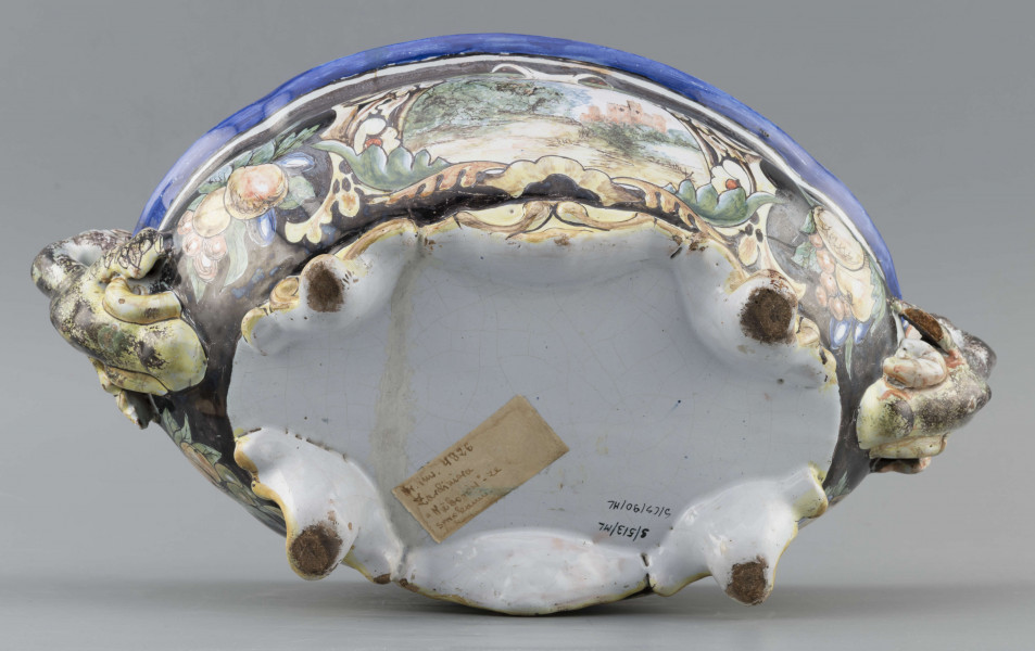 S/CS/90/ML - Żardiniera owalna, na czterech konsolkowych nóżkach połączonych falistymi spływami, wylew z lekko rozchylonym brzegiem, po bokach dwa uchwyty w formie uskrzydlonych smoków, pionowo osadzone. Powierzchni malowana wielobarwnie:  po obu stronach bogate kartusze wolutowo-liściaste z pejzażami  -  jeden z zamkiem obronnym, drugi z wierzbą, po bokach kartuszy festony owoców na ciemno granatowym tle. Wylew obwiedziony błękitnym pasem, wnętrze białobłękitne. 