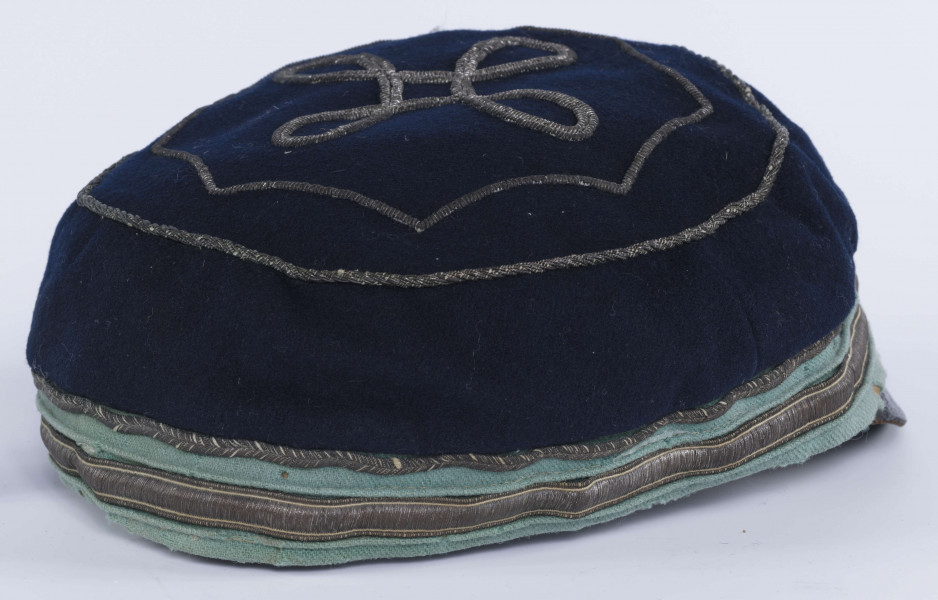 ML/H/2208 - Czapka, czyli tzw. dekiel członka Polskiej Akademickiej Korporacji Astrea działającej na Katolickim Uniwersytecie Lubelskim w latach 1923-1939. Nakrycie głowy w barwach korporanckich, uszyte z nieusztywnianego granatowego sukna, z daszkiem z czarnej skóry, z otokiem w kolorach błękitnym i srebrnym. Na zewnętrznej stronie denka w centralnej części wyszyty fabrycznie srebrną nicią znak korporacji (stylizowany na gwiazdę, w postaci połączonych ze sobą ozdobnych czterech liter „A”) umieszczony wewnątrz haftowanego oktogramu otoczonego kołem z naszytego ozdobnego sznureczka. Po bokach widoczne miejsca zszycia materiału. Wewnątrz podszewka w kolorze granatowym oraz wzmacniający skórzany pasek w kolorze brązowym naokoło.