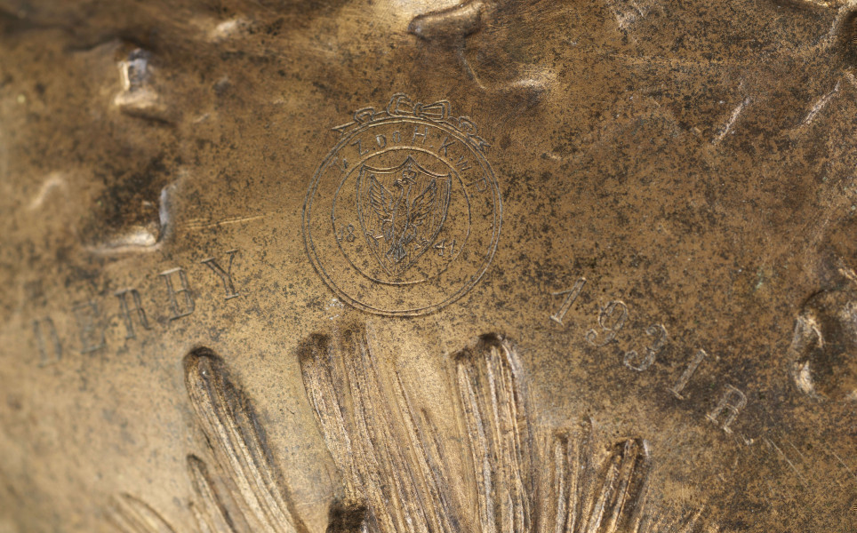 S/Mt/427/ML - Waza w kształcie spłaszczonej kuli, z niską, kolistą stopą i krótką szyją o lekko wywiniętym brzegu. Powierzchnia zdobiona reliefem: po bokach dwa uchwyty w formie głów żubrów, wokół nich gałązki dębu, pośrodku korpusu gałązki sosny z szyszkami wyrastające ze stopy. U góry na brzuścu rytowany napis majuskułą 